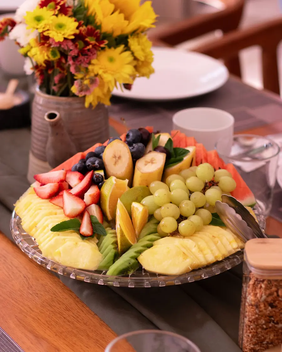 Travessa com morango, abacaxi, uva, banana e kiwi numa mesa de madeira com flores amarelas