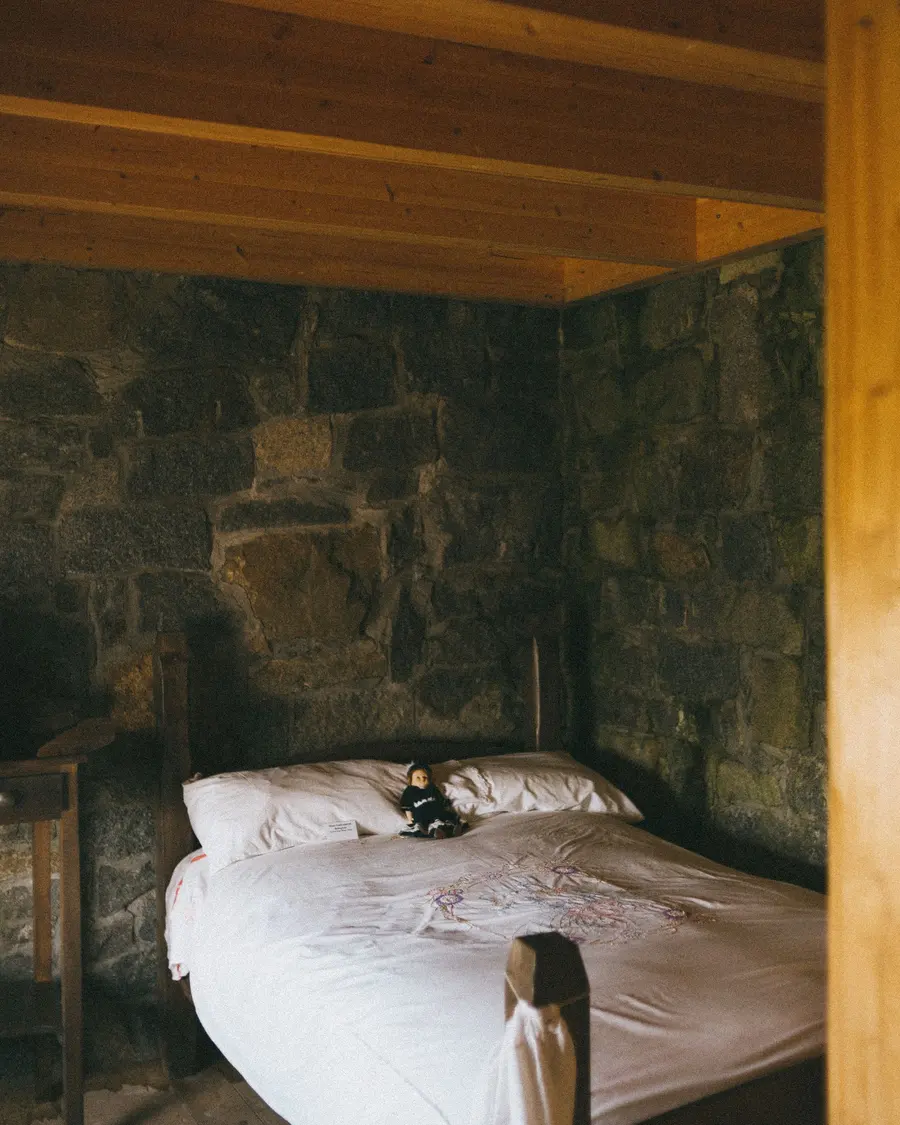 Quarto com parede de pedra, cama de casal com lençóis brancos e teto de madeira